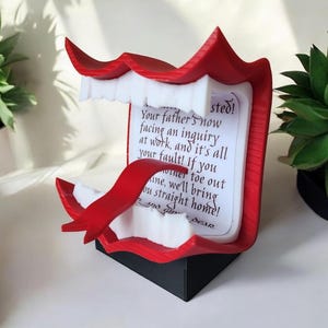 Peut inclure: Une bouche rouge en 3D imprimée avec des dents blanches et un ruban rouge. La bouche est ouverte et une petite carte blanche avec du texte noir est à l'intérieur. Le texte dit : "sted! Your father's now facing an inquiry at work, and it's all your fault! If you oner toe out ane, we'll bring ou straight home! Dear."