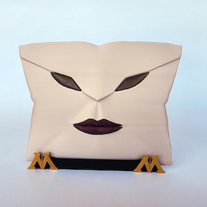 Pode incluir: Uma caixa decorativa bege claro em forma de rosto com formas de olhos castanhos escuros e lábios roxos escuros. A caixa tem uma alça preta e detalhes dourados na base, com um design estilizado "M". O design geral é artístico e único.