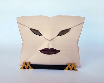 May include: A light beige, face-shaped decorative box with dark brown eye shapes and deep purple lips. The box has a black handle and gold-colored accents at the base, featuring a stylized "M" design. The overall design is artistic and unique.