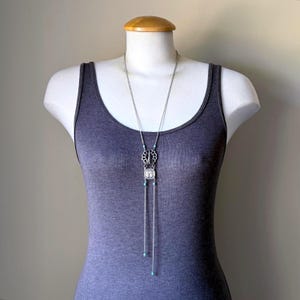May include: Silver-toned necklace with a decorative pendant and turquoise accents. The necklace features a long chain with a geometric design and a square-shaped centerpiece. The necklace is displayed on a mannequin wearing a gray tank top.