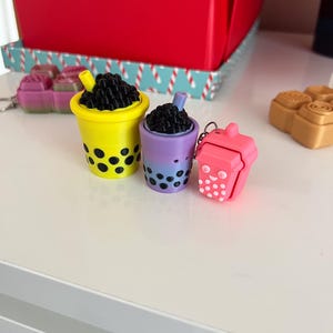 Puede incluir: Tres llaveros de té boba de colores en amarillo, morado y rosa. La taza amarilla tiene boba negra y puntos negros, la taza morada tiene boba negra y puntos negros, y la taza rosa tiene una cara sonriente y puntos blancos. Una caja roja está en el fondo.