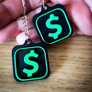 May include: Two black keychains with a green dollar sign symbol. The dollar sign is inside a green square border. The keychains are attached to a silver chain and ring. The background is a wooden surface.