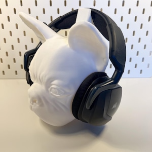 Könnte beinhalten: Schwarze Over-Ear-Kopfhörer mit einem weißen 3D-gedruckten Bulldoggenkopf-Ständer. Die Kopfhörer haben einen schwarzen Kopfbügel und Ohrmuscheln mit einem silbernen Logo.