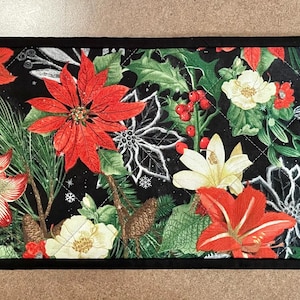 May include: A rectangular, quilted Christmas table runner with a black border. The runner features a floral pattern with red poinsettias, white and red amaryllis, white sketched flowers, and green foliage on a black background.