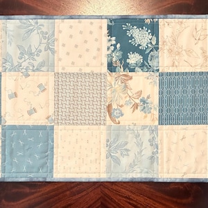 May include: A rectangular quilted table runner featuring a patchwork design. The runner is composed of squares in shades of blue, cream, and beige, with floral and geometric patterns. A blue border frames the runner, adding a cohesive finish.