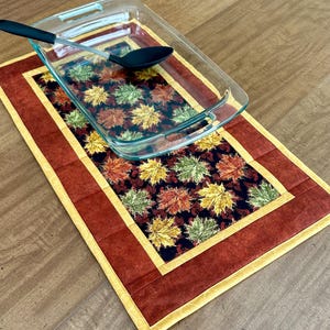 May include: A clear glass baking dish and black spoon rest on a fall-themed table runner. The runner features a dark background with colorful autumn leaves and a red border with a yellow trim. The spoon is approximately 12 inches long.