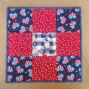 May include: A red, white, and blue patchwork quilt with a gnome pattern in the center. The quilt is made of squares with different patterns, including stars, hearts, and gingham.