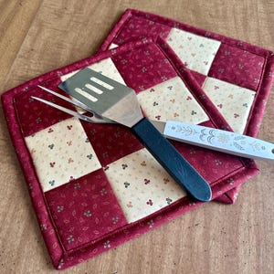 May include: Two quilted pot holders with a patchwork design in burgundy and cream, featuring a floral pattern. A stainless steel spatula and a serving fork with black and white handles rest on top, suggesting kitchen or dining use.