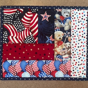 May include: A patriotic quilted placemat featuring a patchwork design with red, white, and blue fabrics. The design includes stars, stripes, and teddy bears, with a navy blue border. The placemat measures approximately 12 x 16 inches.