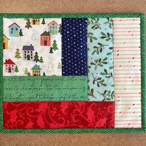 May include: A rectangular Christmas-themed quilted placemat. The placemat features various fabric patterns, including houses, trees, polka dots, mistletoe, and red swirls. The border is green, and the text "all the way joyous noel the west merry christmas tis the season" is visible.