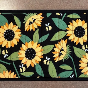 May include: A rectangular quilted table runner featuring three distinct sections. The outer panels are light yellow with black cursive text and small bee illustrations. The center panel displays a black background with yellow sunflowers and bees.