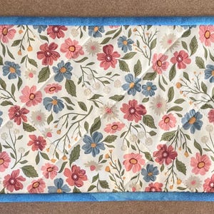 May include: Rectangular floral table runner with a light blue border. The runner features a pattern of pink, blue, and white flowers with green leaves on a cream background. The runner is approximately 24 inches long and 12 inches wide.