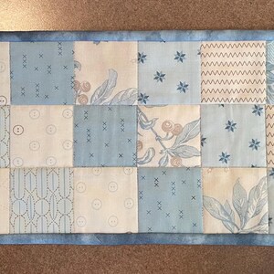 May include: A rectangular patchwork table runner featuring a variety of light blue and cream-colored squares. The squares display floral, geometric, and button patterns. The runner is bordered with a solid light blue fabric.