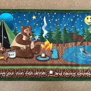 May include: A rectangular quilted fabric wall hanging featuring a nighttime camping scene with a bear, a tent, a campfire, and a pond. The bottom border includes the text: moonlit nights, making your own fish dinner, and having s'mores for dessert.