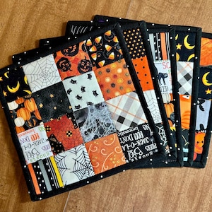 May include: A set of four square fabric potholders featuring a patchwork design with various Halloween-themed patterns including pumpkins, spider webs, black cats, candy corn, and text reading "BOO!" and "JACK-O-LANTERN" with black binding.