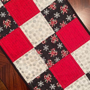 May include: A quilted Christmas table runner with a patchwork design. The squares alternate between red, black with candy cane and snowflake patterns, and silver with snowflake patterns. The runner has a black border.