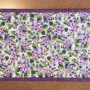 May include: Rectangular table runner featuring a floral pattern of purple hydrangeas on a light blue background. The runner is bordered with a solid purple fabric. This decorative piece is suitable for a dining table or a console.