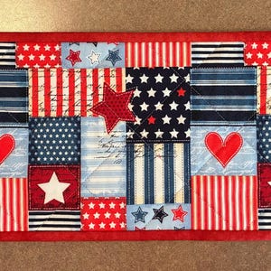 May include: A rectangular patriotic table runner with a red border. The design features alternating blocks of red and white stripes, navy blue stars, and white stars on a blue background. Red hearts and stars are appliqued.