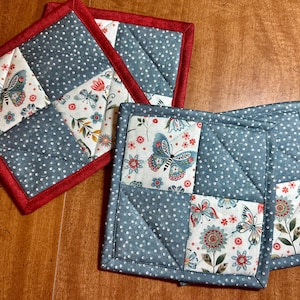 May include: A set of four square quilted pot holders. The pot holders feature a patchwork design with blue polka dot fabric and butterfly and floral print squares. The edges are trimmed with red fabric.