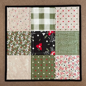 May include: A square, quilted patchwork featuring nine fabric squares with a black border. The squares display various patterns, including stars, polka dots, floral designs, and a green and white gingham check. Colors include beige, green, red, and black.