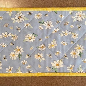 May include: A rectangular quilted placemat with a light blue background, featuring a pattern of white daisies and small bees. The placemat is bordered with a bright yellow trim. The dimensions are approximately 18 inches long and 8 inches wide.