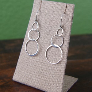 May include: A pair of silver dangle earrings with three interlocking circles. The earrings are displayed on a beige fabric stand.