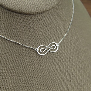 May include: A silver infinity symbol necklace on a silver chain. The necklace is displayed on a brown fabric mannequin.