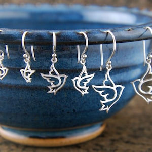 May include: A set of six silver dove earrings hanging from the edge of a blue ceramic bowl. The doves are cut out of the metal and have a delicate, intricate design.