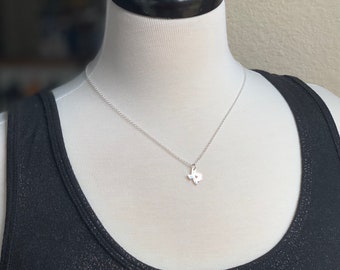 May include: A silver necklace with a small Texas-shaped pendant. The necklace is on a white mannequin wearing a black tank top.