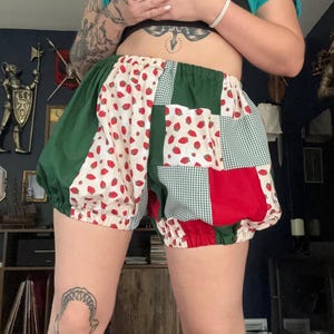 May include: Patchwork bloomers featuring a mix of fabrics including a white strawberry print, solid dark green panels, red fabric, and a green and white gingham check pattern. The shorts have an elasticized waistband and elasticized leg openings.
