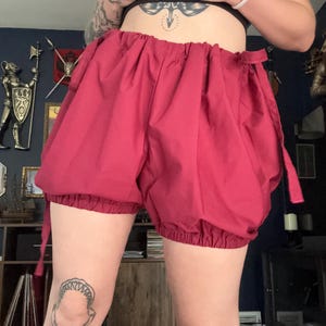 May include: Deep red bloomers featuring a high-waisted design with an elasticized waistband and gathered leg openings. The shorts have a voluminous, puffed silhouette and include thin fabric ties on the sides for an adjustable fit.
