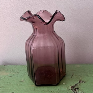 May include: A vintage, amethyst-colored glass vase with a ruffled rim. The vase has a hexagonal base and vertical ribbed detailing. The vase is displayed on a distressed, green-painted wooden surface against a neutral wall.