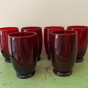 Peut inclure: Six verres à boire rouge rubis. Les verres sont en verre rouge foncé et ont une forme arrondie. Ils sont disposés en groupe sur une surface verte, avec un fond neutre.