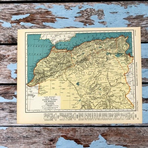Puede incluir: Un mapa antiguo de Argelia, Túnez y Marruecos, con información geográfica detallada. El mapa tiene un fondo crema con texto en negro y rojo. El título "RAND MONALLY" es visible. El mapa está sobre una superficie de madera desgastada azul y marrón.
