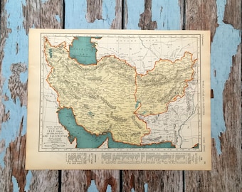 May include: A vintage map depicting Iran, Afghanistan, and Baluchistan, with detailed geographical features. The map has a beige background with green and blue accents, set against a distressed, blue-painted wooden surface.