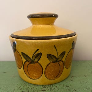 May include: A yellow ceramic jar with a lid. The jar is decorated with orange fruit designs and dark brown accents. The jar is sitting on a green surface.