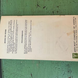 May include: A vintage pamphlet titled "Books on Herbs" with text including "Recipe Magic" and "Jiffy-Pot Company of America." The pamphlet is cream-colored with black text and a green logo, lying on a green wooden surface.