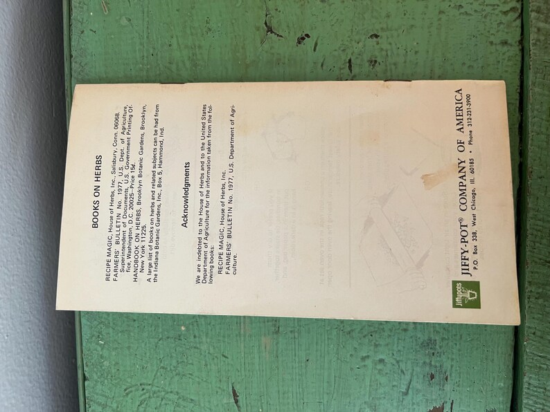 May include: A vintage pamphlet titled "Books on Herbs" with text including "Recipe Magic" and "Jiffy-Pot Company of America." The pamphlet is cream-colored with black text and a green logo, lying on a green wooden surface.