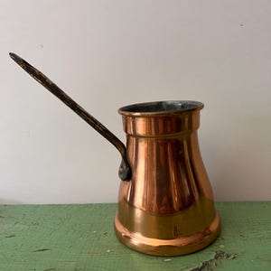 May include: A copper coffee pot with a long, dark handle. The pot has a rounded base and a flared top. The copper has a warm, shiny finish. The handle is attached with a small, dark metal piece. The pot is likely used for making Turkish coffee.