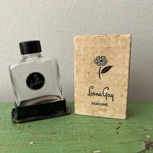 May include: Vintage perfume bottle with a black cap and a black label that reads "Lorna Gay". The bottle sits on a black base next to a small, rectangular box with the words "Lorna Gay PERFUME" and a flower illustration.