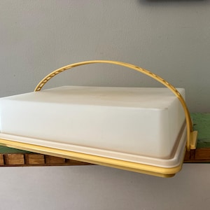 Puede incluir: Un portatartas rectangular blanco con asa y ribete amarillos. El portatartas tiene tapa y base, y el asa se arquea sobre la parte superior. El portatartas está sobre una superficie verde y marrón.