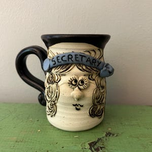 Könnte beinhalten: Eine handgefertigte Keramiktasse mit Gesichtsdesign. Die Tasse ist cremefarben mit schwarzen Akzenten und einem schwarzen Griff. Ein blaues Banner oben zeigt "SECRETARY". Das Gesicht hat große Augen, eine kleine Nase und einen lächelnden Mund.