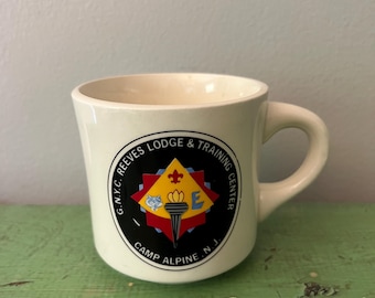 Puede incluir: Taza de cerámica color crema con asa curva. La taza presenta un emblema circular con el texto "G.N.Y.C. Reeves Lodge & Training Center Camp Alpine N.J.". El emblema incluye una antorcha, una flor de lis y la letra "E".