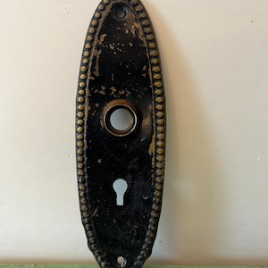 Puede incluir: Placa de puerta ovalada negra antigua con un ojo de cerradura y una abertura circular para el pomo. La placa está bordeada con un borde decorativo de cuentas doradas en relieve. La pintura está desgastada, mostrando signos de edad.
