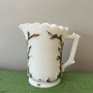 May include: A white milk glass creamer with a scalloped rim and a handle. The creamer is decorated with gold-colored floral designs. The creamer sits on a small, round base. The background is a light green surface.