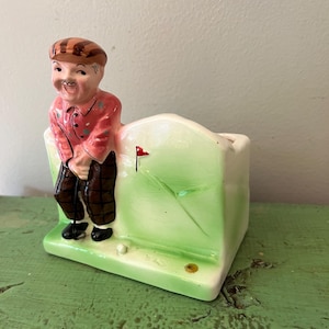 May include: A vintage ceramic planter featuring a golfer figure leaning against a green golf course scene. The golfer wears a red shirt, brown plaid pants, and a brown cap. A red flag marks the hole.