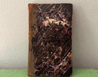 Op de afbeelding: Een klein antiek boek met een gemarmerde kaft in bruin, zwart en rood. De rug is lichtbruin. Het boek ligt op een verweerd, groen geverfd oppervlak. De kaft van het boek vertoont slijtage.