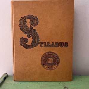 May include: A vintage tan-colored book titled "SYLLABUS" with an ornate "S" design and a university seal. The book's cover features the text in a dark brown color. The university seal includes the text "NORTHWESTERN UNIVERSITY" and the year "1851".