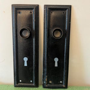 Op de afbeelding: Twee rechthoekige, zwarte deurplaten met een sleutelgat en een cirkelvormige opening voor de deurknop. De platen hebben een licht getextureerd oppervlak.