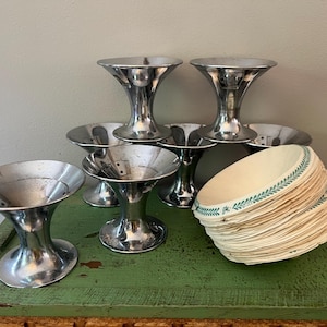 May include: A collection of vintage metal ice cream sundae cups with a flared design. A stack of paper liners with a green floral border sits nearby. The items are arranged on a green surface.